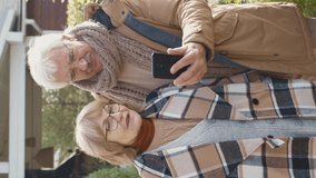 Vertical view shot of senior Caucasian woman and her husband wearing warm casual clothes and eyeglasses standing in garden near house and having video call using telephone