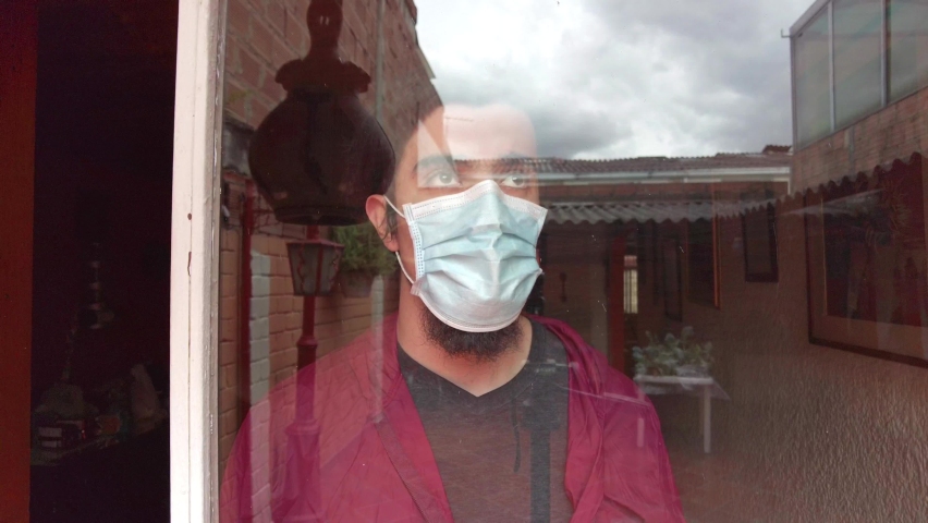 Young man in quarantine, with face mask and red jacket looking out the window with his hands on it. Sad  worried expression. 
Window reflecting house roofs and sky. COVID or Coronavirus pandemic scene