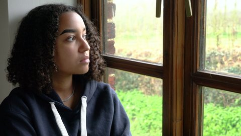 Beautiful mixed race biracial African American girl teenager young woman wearing a blue hoodie sitting looking out of a window looking sad then happy