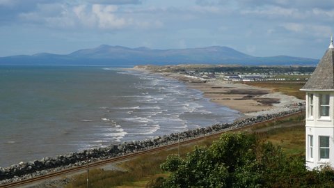 Looking past an old building, across railway line through to Coastal waves to beach and in the background can be seen the mountains of the Llyn Peninsula, Wales, UK - 10 Second version