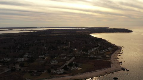 an aerial view over the eastern end of Orient Point, Long Island during sunset. The camera dolly in and truck right and tilt down as the sunsets in the cloudy sky, over the quiet seaside neighborhood.