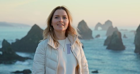 Happy young woman enjoying wilderness nature landscape view. Attractive female traveler portrait in soft sunset light. Slow motion smiling model with scenic rocky sea cliffs on background. Footage 4K