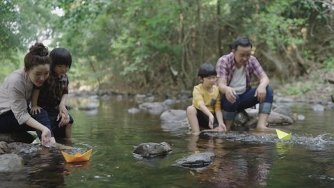 An Asian family, a father and a mother teach their two sons kids  how to craft a paper boat. Float in a natural forest stream. Joyfully and happily On vacation. Children's learning concept.