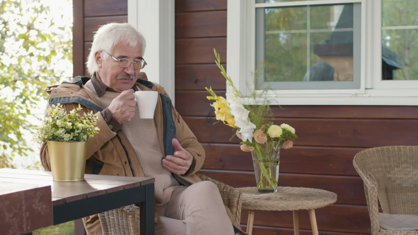 Medium shot of senior Caucasian man wearing warm clothes and eyeglasses sitting on terrace and drinking tea. Then his teenage grandson brining him present and he hugging him