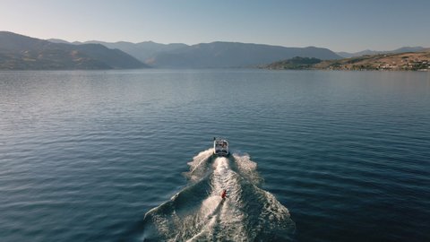 Drone Following Boat Towing Wakeboarder on Scenic Lake Chelan with Desert Mountains. Aerial view of man riding board behind speedboat on scenic desert body of water