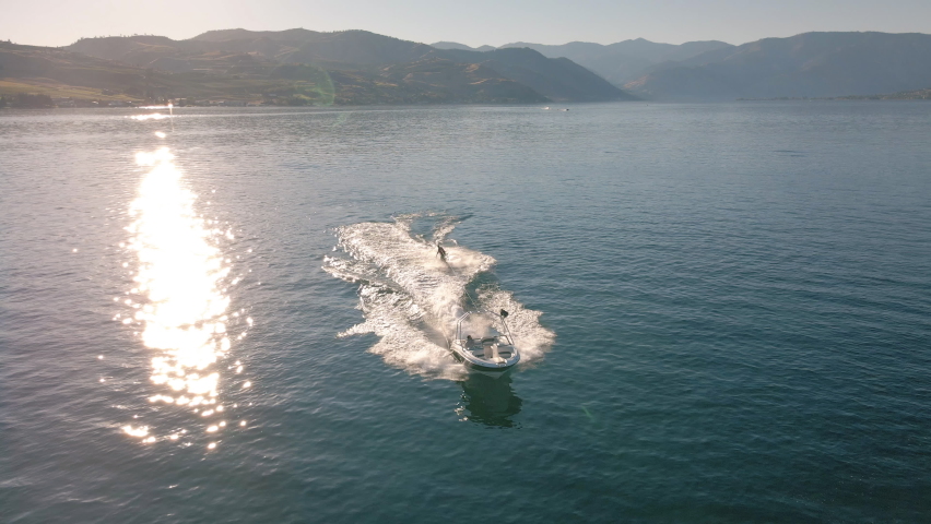 Drone Flying Over Boat Towing Wakeboarder Spraying Water on Sunny Summer Day. Sunshine twinkle off scenic lake water with man riding board behind speedboat on Lake Chelan Royalty-Free Stock Footage #1069999237