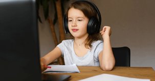 Cute little girl with headphones using laptop to study at home, writing, answer, online learning, education, social distance, quarantine coronavirus.