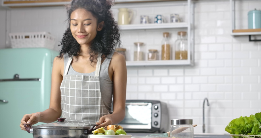 A young, happy and beautiful Asian woman with curly long hair wearing grey gingham apron. She's making a soup for breakfast and open a pot for smelling, scooping up with a smile in the modern kitchen. | Shutterstock HD Video #1070000740