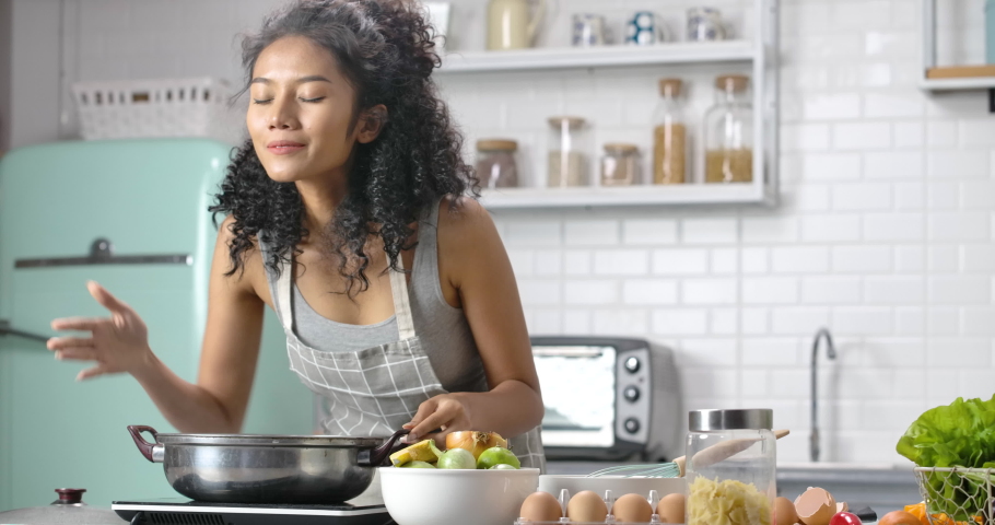 A young, happy and beautiful Asian woman with curly long hair wearing grey gingham apron. She's making a soup for breakfast and open a pot for smelling, scooping up with a smile in the modern kitchen. Royalty-Free Stock Footage #1070000740