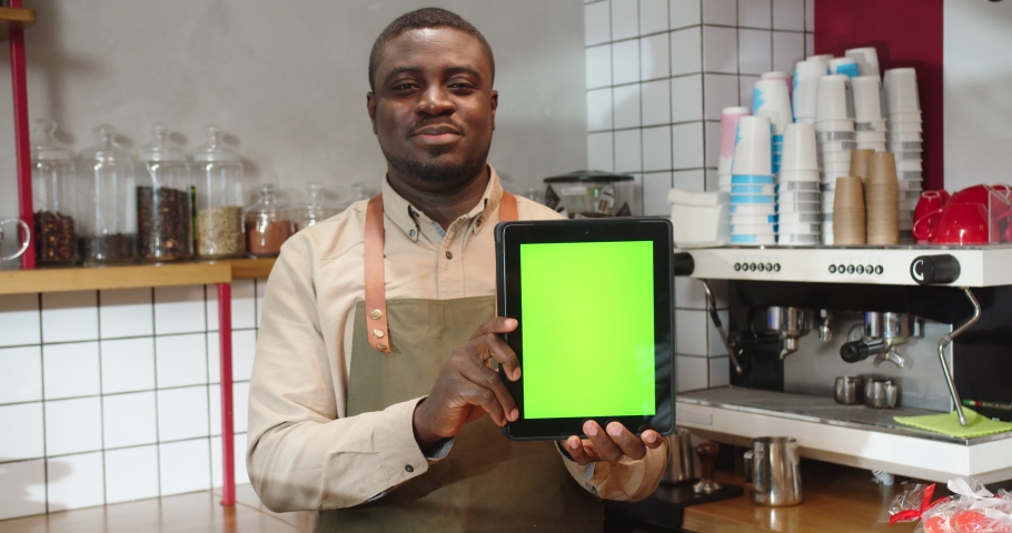 Attractive male barista holding digital tablet with green screen in coffee shop. Adult Afro American man showing chroma key on gadget. Small business, entreprenuership concept.