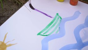 Closeup view video footage of little boy of 5 years old painting outdoors together with his mommy. Child paints blue sea water, sun, beach, sailboat on water on sheet of white paper laying on ground