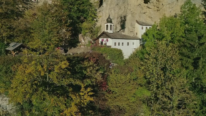 An aerial drone shot of a secluded white structure with a bell tower built into the side of a rocky cliff face - Daytime.