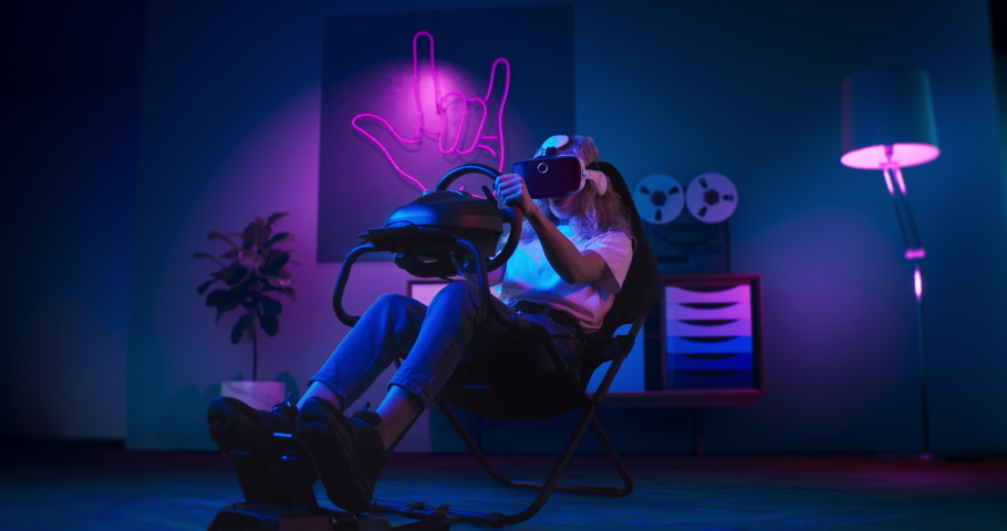 A girl is playing racing game in VR. She is sitting on the gaming chair with a steering wheel and pedals. A girl wears VR headset. Chair is located in the living room with red and blue neon lighting. Royalty-Free Stock Footage #1070012770