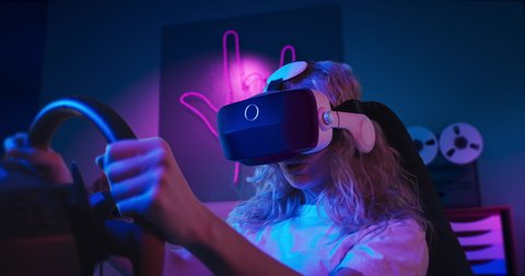 A girl is playing racing game in VR. She is sitting on the gaming chair with a steering wheel and pedals. A girl wears VR headset. Chair is located in the living room with red and blue neon lighting.