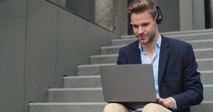 Handsome Businessman works distantly, sits on Stairs near Office Building. Young Male is using Laptop and headset for Communication with his multinational Business Partners outdoor. Webinar. Online.