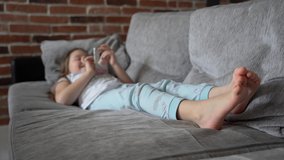A little girl lies at home on the couch and looks into a smartphone. The child plays and watches the video. The girl has bare feet. The child is wearing pajamas. 4K
