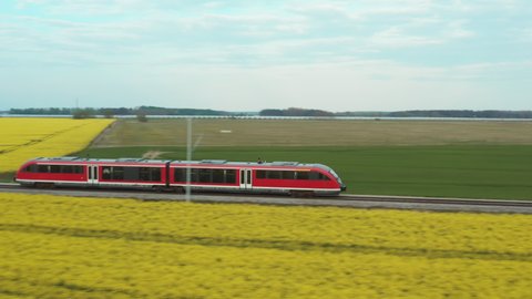 A high-speed red electric train rushes through a blooming field of rapeseed. Ecological transport in front of a source of alternative biofuel.