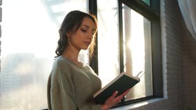 Beautiful woman sits near window and reads a book on background of sunset.