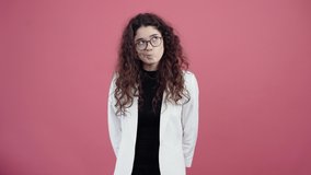 The intelligent young woman with curly hair is thinking of new ideas and strategies for solving a problem. Young hipster in white jacket and black shirt, with glasses posing isolated on pink