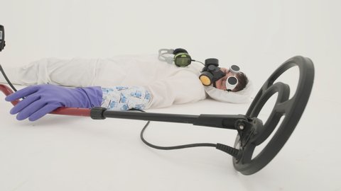 Close-up of man in white chemical protection suit, gas mask and purple rubber gloves lies on the floor. There is headfones on his chest. His hand is on the metal detector which lies near him. Filmed 