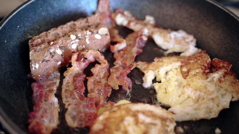 Top view of fatty breakfast on sizzling pan, process of english breakfast or lunch preparation at home. High-calorie food frying on gas stove, crispy bacon strips, eggs and a slice of rye bread