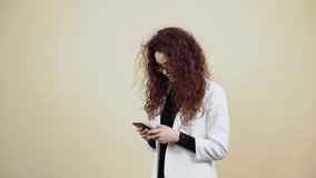 Happy young woman with curly hair holds a phone in her hand and vb with friends through the message. Young hipster in gray jacket and white shirt, with glasses posing isolated on beige background in