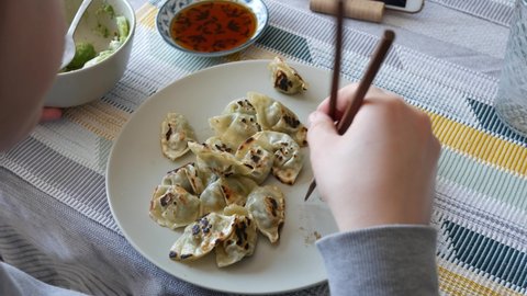 A woman is sitting at her home eating dumplings Gyoza with soy sauce using chopsticks