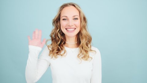 A beautiful woman is waving her hand to the camera doing hello gesture while standing isolated over blue wall in a studio