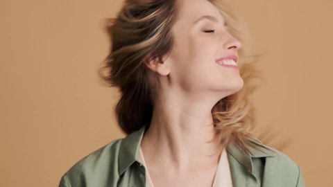 A close-up view of a happy young woman is shaking her hair standing isolated over beige background in studio