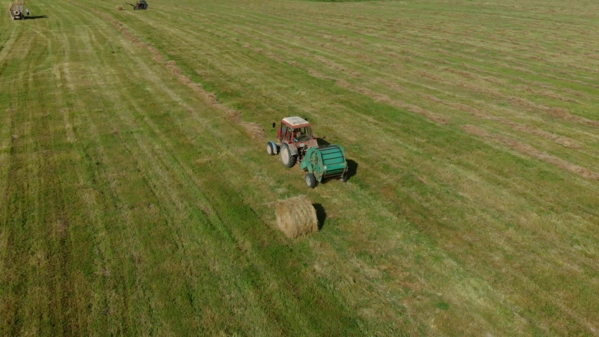 Harvesting hay. A farmer on tractor with round baler rides on wheat field after harvesting and makes hay bales. Agricultural machinery collects dry straw in rolls and round large bales. Aerial view 