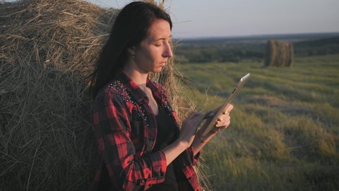 Smart technologies in agriculture. A pretty young woman farmer uses a tablet near a haystack in a mown field at sunset, planning and running an agribusiness using smart technologies.