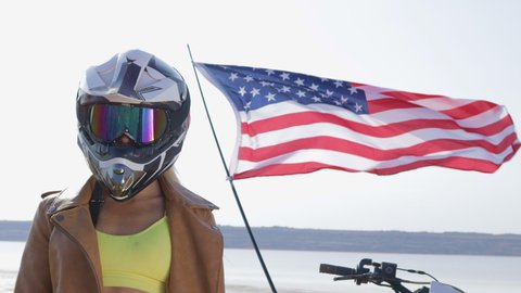 A portrait of a girl standing in a protective motorcycle helmet against the background of the USA flag waving in the wind.