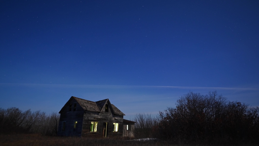 Time lapse of stars moving slowly through the night sky above a dilapidated house. Light can be seen through the broken windows.
