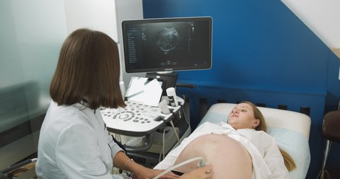 Pregnant woman getting ultrasound sonogram scan, while young concentrated female doctor obstetrician explains procedure to her. Pointing At The Screen, Showing The Baby's Ultrasonic Image.