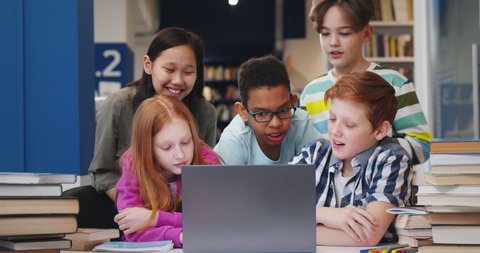 Happy multiethnic classmates using computer together in school library. Portrait of smiling diverse schoolchildren studying on laptop at desk in library