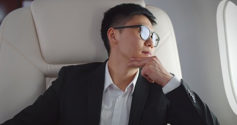 Thoughtful asian businessman on airplane looking outside window. Portrait of stressed korean entrepreneur putting off eyeglasses relaxing in leather chair flying first class