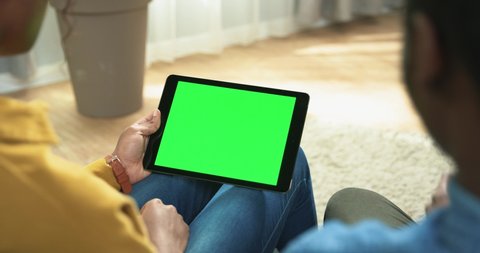 Close up of couple man and woman tapping on tablet with chroma key sitting in room at home. Over shoulder view of woman typing on device with green screen searching internet, tech concept