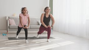 Active sports training. Cheerful african american young mother and her teen daughter exercising forward lunges at home together, tracking shot, slow motion
