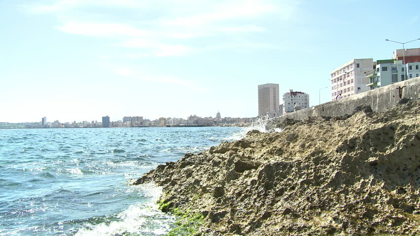 A wide shot with waves crashing into rocks in the foreground and a landscape view of the city of Havana, Cuba in the background