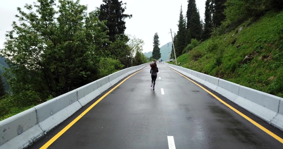 A girl runs on wet asphalt in the mountains. All around are green trees and grass, forest. Light rain and cloudy weather. The girl goes in for sports. Puddles on the asphalt. Cloudy sky. Fresh air.