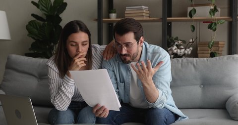Young stressed married couple sit on couch hold papers reading bad news in legal document, got warning notice feel shocked looking frustrated. Pay penalty, unpaid debt, eviction notification concept
