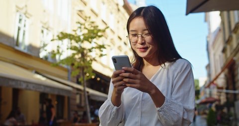 Happy female student in glasses standing outside cafe and using smartphone. Young Asian woman reading messages, texting friends, looking around and smiling in city street. Communication concept.