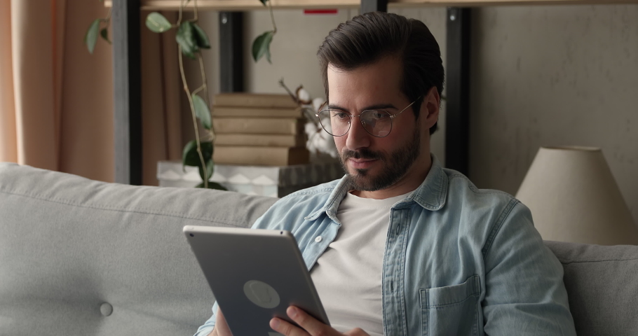 Busy attractive man in glasses sit on sofa holding tablet device. Do telecommute job remotely, chatting in social media, choose goods on web stores. E-commerce, virtual services, fun app user concept Royalty-Free Stock Footage #1070068915