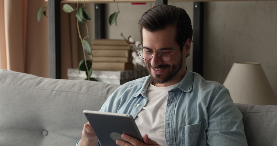 Busy attractive man in glasses sit on sofa holding tablet device. Do telecommute job remotely, chatting in social media, choose goods on web stores. E-commerce, virtual services, fun app user concept | Shutterstock HD Video #1070068915