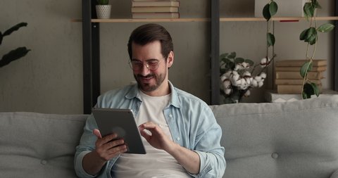 Happy young single man relax alone at home sit on sofa hold digital tablet enjoy e-date services, surfing web, studying online, shopping on internet use social media apps. Modern device usage concept