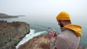 A man standing on the cliff edge is making video with drone of beautiful marine landscape and sea waves crashing with foam.