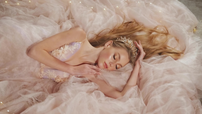 Fantasy blonde girl princess in golden crown. Medieval woman in vintage long pink peach bridal dress. queen lies in lush tule fabric of fluffy voluminous skirt. sleeping Beauty Fashion model dreaming | Shutterstock HD Video #1070070040