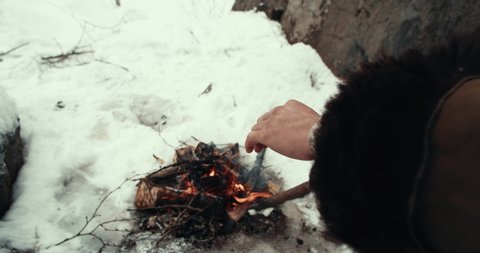 A man wanderer in a warm sheepskin coat warms his hands in winter by a burning fire near the rocks.