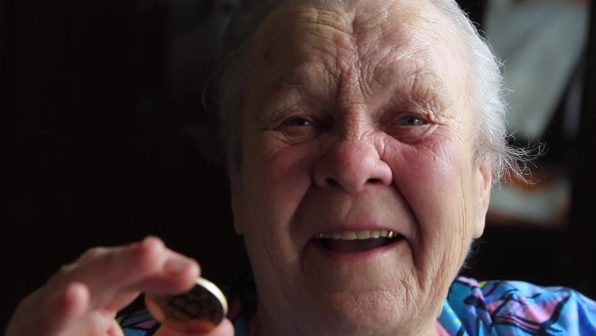 An elderly woman, a grandmother, is holding a bitcoin coin in her hands and leans it to her eye
