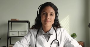 Concentrated skilled young female physician doctor wearing headphones with microphone, giving online virtual consultation to patient or taking part in video conference, medical service concept.